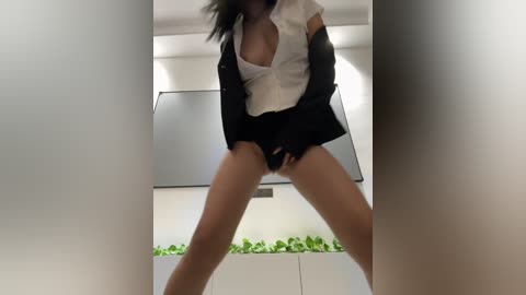 Media: Video of a woman with light brown skin, wearing a white blouse unbuttoned to reveal cleavage, black blazer, and black shorts, standing with legs apart in a modern, minimalist room with green plants.