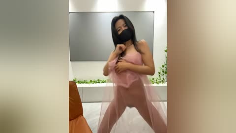 Media: Video of an Asian woman with black hair, wearing a pink sheer dress, black mask, and black gloves, in a modern room with a large mirror and green plants.