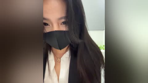 Media: Video of a masked woman with long black hair, wearing a white shirt and black blazer, partially obscured by a blurred, shadowy figure.