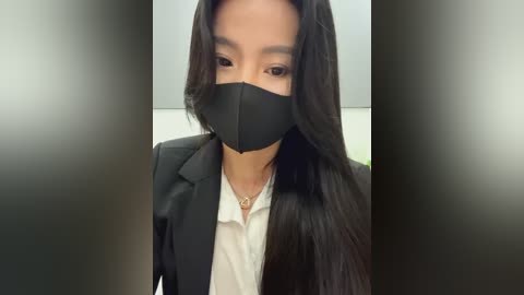 Media: Video of an Asian woman with long black hair, wearing a black face mask and a black blazer over a white shirt, standing indoors with blurred background.