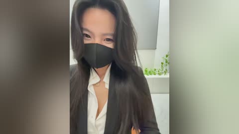 Media: Video of a woman with long black hair, wearing a black face mask, black blazer, and white shirt, standing in a modern bathroom with white tiles and a green plant.