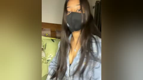 Media: Video of a young Black woman with long, straight black hair, wearing a black mask, denim jacket, and a necklace, in a dimly lit room with green and beige walls.