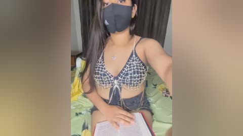 Media: A video of a young woman with long black hair, wearing a black face mask and a black and white checkered bra, seated on a bed with a green floral duvet, reading a book.