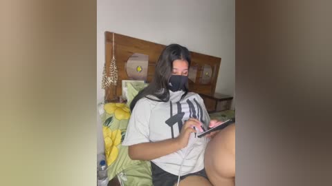 Media: Video of a young woman with medium skin tone, wearing a black mask, grey T-shirt, and black shorts, sitting on a bed, reading a book, surrounded by colorful bedding and wooden headboard.