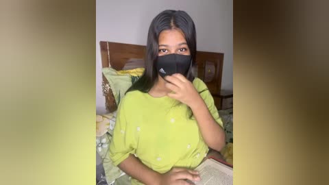 Media: Video of a young South Asian woman with medium-brown skin, wearing a green top and black mask, sitting on a bed with patterned bedding.