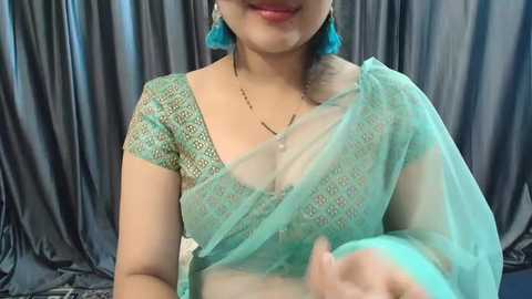 Media: Video of a smiling South Asian woman with fair skin, wearing a sheer green sari with lace detailing, turquoise earrings, and a gold necklace. Background features dark gray, draped fabric.