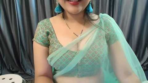 Media: Video of a smiling South Asian woman with light skin, wearing a teal lace blouse and matching saree. She has turquoise earrings and a necklace. Background is a dark gray curtain.