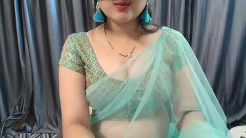 Media: Video of a South Asian woman with light brown skin, wearing a teal saree with intricate lace, turquoise earrings, and a gold necklace. She stands against grey satin curtains, her face partially visible.