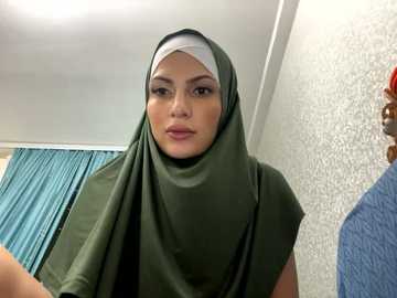 Media: Video of a young woman with fair skin, wearing a dark green hijab and white headscarf, standing indoors with textured white wall, blue curtains, and a partially visible blue shirt.