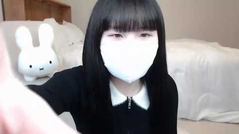 Media: A video of an Asian woman with long black hair and a white mask, standing in a minimalist bedroom with white bedding and a bunny plush toy.