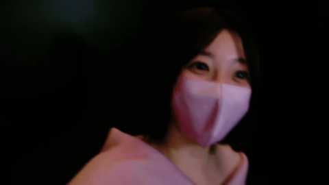 Media: Video of an Asian woman with short black hair and light skin, wearing a pink face mask, black top, and a dark background.