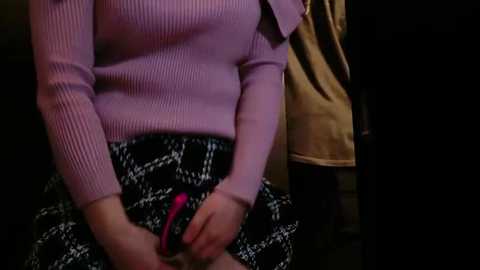 Media: Video of a person in a light purple sweater with long sleeves, holding a pink pen. They are seated, wearing a black and white checkered skirt. Background features a beige garment and dark setting.