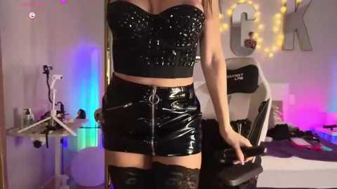 Media: Video of a woman in a black, strapless, sequined top and matching leather mini skirt, sitting in a gaming chair, surrounded by colorful LED lights, and a \"LOL\" sign on a wall.