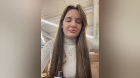 Media: Video of a young woman with long, straight brown hair, wearing a white turtleneck sweater, and a silver necklace, standing in an airport terminal with beige walls and a high ceiling.