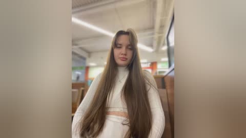 Media: Video of a young woman with long brown hair, wearing a white sweater and skirt, seated in a dimly lit, modern indoor setting with fluorescent lights and wooden chairs in the background.