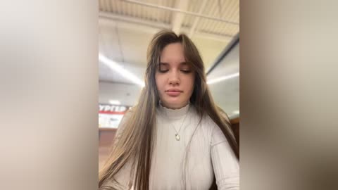 Media: Video of a young woman with long, straight brown hair, wearing a white turtleneck sweater, standing in a dimly lit room with fluorescent lights and blurred signs.