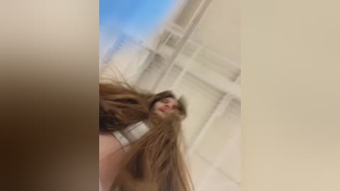 Media: A video of a young woman with long, straight brown hair, captured in motion with a slight blur. She has light skin and is wearing a white top. The background features a white wall and a blue sky.