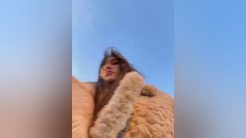 Media: A video of a young Asian woman with long, wavy brown hair, wearing a fluffy, light beige fur coat, against a clear blue sky.