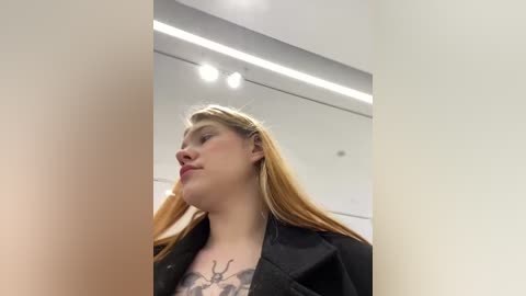 Media: Video of a young Caucasian woman with long blonde hair, wearing a black jacket, standing indoors under bright fluorescent lights. She has a tattoo of a deer's head on her chest.