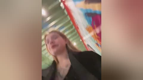 Media: Blurred video of a blonde woman with long hair, wearing a black jacket, walking in a modern indoor space with colorful, abstract art on the wall.