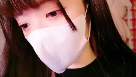 Media: Video of a young East Asian woman with long black hair, wearing a white surgical mask, looking directly at the camera. Background features a red and white checkered pattern.