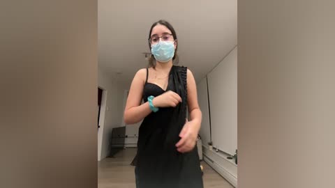 Media: A video of a young woman in a black dress, wearing a blue face mask, standing in a minimalist, beige-walled room. She appears to be adjusting her dress.