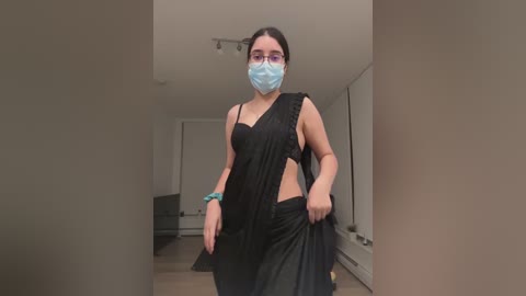 Media: A video of a woman in a black saree and blue face mask, holding her skirt, in a modern, minimalist living room with white walls and wooden flooring.