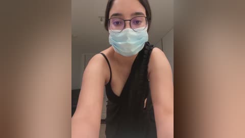 Media: A video of a young Asian woman with glasses, wearing a black lace dress and a blue face mask, standing in a hallway with beige walls.