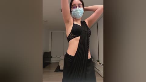 Media: Video of a woman with light skin and short dark hair, wearing a black lace bra and pants, a face mask, and glasses, standing in a modern, minimalist living room.