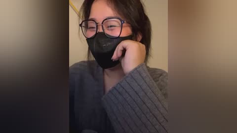 Media: A video of an Asian woman with short, dark hair, wearing glasses, a black mask, and a gray ribbed sweater, leaning on a wall, eyes closed.