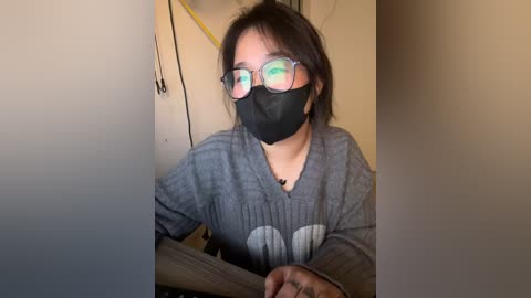 Media: Video of an Asian woman with short, dark hair wearing a gray long-sleeve shirt, black mask, and rectangular glasses, seated indoors with a beige wall and closed door background.