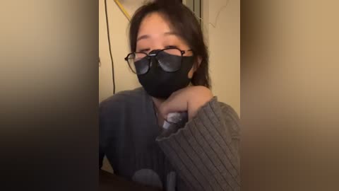 Media: Video of an Asian woman with light skin, wearing black face mask, round glasses, and a gray sweater, leaning against a beige wall, taken indoors.