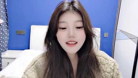 Media: Video of an Asian woman with long black hair, wearing a beige fuzzy robe, sitting on a white bed in a modern bedroom with blue walls and white furniture.