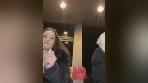 Media: A video of a young woman licking an ice cream cone, taken indoors with dim lighting and a blurred background. She is wearing a black coat.