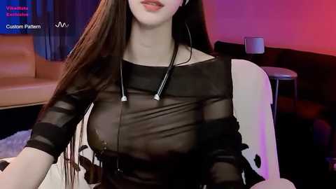 Media: Video of a fair-skinned woman with long, straight brown hair, wearing a sheer black top revealing her large breasts and nipples, seated in a modern living room with red and blue lighting.