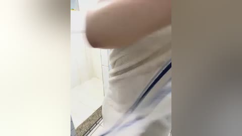 Media: A video of a person in a white shirt and light blue pants, taken from an angled perspective, shows a blurred arm and partially visible face. The background features a tiled bathroom with a shower curtain.