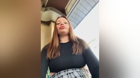 Media: Video of a plus-sized woman with fair skin, long brown hair, wearing a black long-sleeved top and plaid skirt, standing in a hallway with beige walls and a white ceiling.