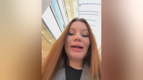 Media: Video of a young Caucasian woman with long, straight, light brown hair, wearing a gray sweater over a black top. She's smiling with her mouth open, standing in a modern, brightly lit atrium with glass walls and circular railings.