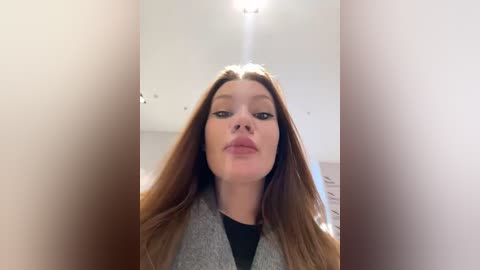 Media: A video of a fair-skinned woman with long, straight, light brown hair, wearing a gray cardigan over a black top, indoors with a white ceiling and soft lighting.