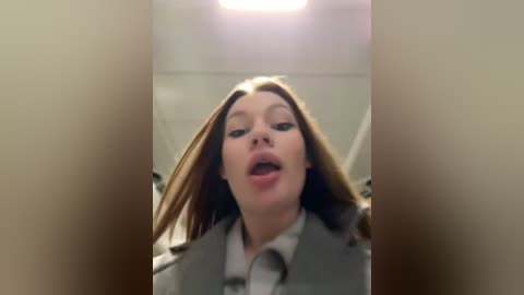 Media: Video of a woman with long, straight brown hair, wearing a gray coat, mouth open in apparent surprise, blurred background of an indoor setting with fluorescent lighting.