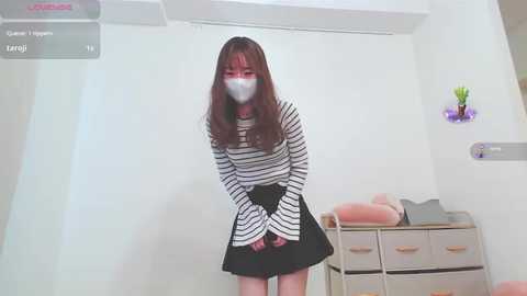 Media: Video of an East Asian woman with long, straight brown hair, wearing a striped top and black skirt, standing in a minimalist room with white walls, a desk, and a potted plant.