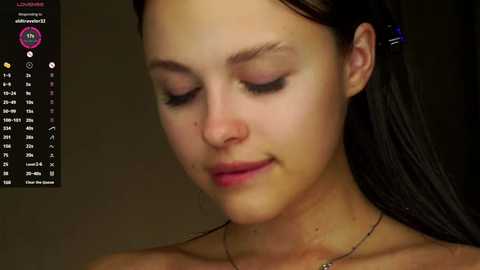 Media: A video of a young woman with fair skin, long dark hair, closed eyes, and a subtle smile. She wears a black top and a thin necklace. The background is dark and indistinct.