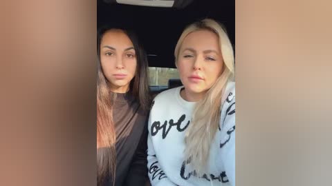 Media: Video of two women, one with long, straight, dark hair and the other with long, straight, blonde hair, wearing black and white \"love\" sweatshirts, sitting closely in a car.