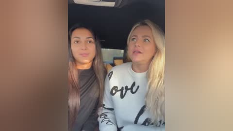 Media: Video of two women in a car. The woman on the left has long dark hair, wearing a black sweater, and the woman on the right has long blonde hair, wearing a white sweater with \"Love\" written on it.