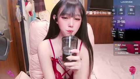 Media: Video of an East Asian woman with long black hair and fair skin, wearing red lingerie, drinking from a glass, in a cozy bedroom with wooden furniture and a TV showing a gaming stream.