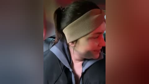 Media: Video of a woman with long dark hair tied back, wearing a beige headband, and a dark jacket, seated in a car with a red light illuminating her face.