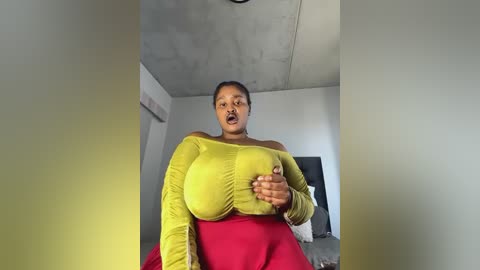 Media: A video of a plus-size Black woman with a medium complexion, wearing a bright yellow off-shoulder top and red skirt, seated indoors with a surprised expression, hands over her chest.