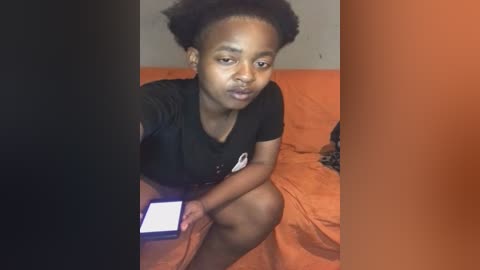 Video of a young Black woman with short natural hair, sitting on an orange bed, wearing a black shirt, holding a smartphone, in a dimly lit room with a dark vertical partition.