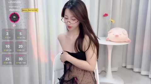 Media: A video of an East Asian woman with long black hair, wearing glasses, a black off-shoulder top, and holding a black polka-dot skirt. She sits in a minimalist, white room with a small pink pig statue on a white table.