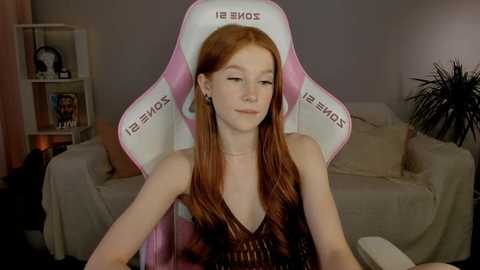 Media: Video of a fair-skinned, red-haired woman with long hair, wearing a black, sleeveless top, sitting in a pink \"Sony\" gaming chair in a cozy living room with beige furniture and plants.